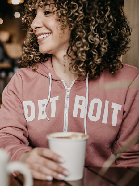 Detroit Classic full zip hoodie by Pure Detroit in mauve color with Detroit printed in white across the chest and worn by an attractive female smiling and drinking coffee