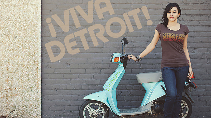 Woman standing next to a teal scooter against a wall with 'Viva Detroit!' graffiti and wearing a Detroit Soul T-shirt by Pure Detroit